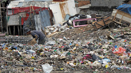 A man bends over and rummages through piles of garbage that surround him.
