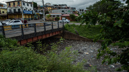 Garbage collects in a stream under a bridge. In the background, cars are parked in front of shops and homes.