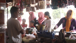 An Indonesia market trader stands at his stall while women customers look at the food on sale