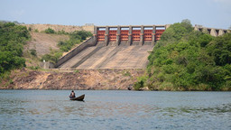One man in a small boat on a reservoir, in front of a large dam