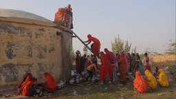 A group of women use a ladder to climb up to an imposing well to collect water in a desert-like landscape