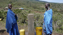 Two women stand on a hillside next to a concrete post with two taps, waiting for their water containers to fill.