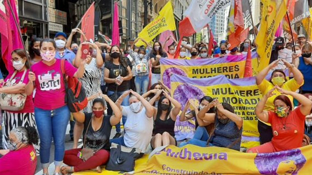 Women wearing masks and carrying banners make a triangle sign with their arms.