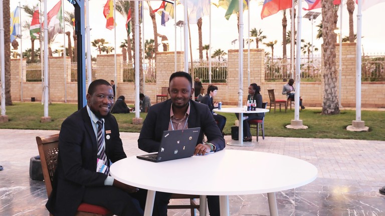 Olivier Ishimwe and Yamikani Idriss sit on a table with world flags in the background