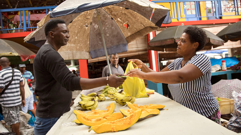 A woman market trader hands over a bag of produce to a male customer, who is giving her money in return