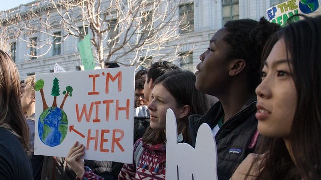 A group of young people holding a poster that reads "I'm with her" - the poster shows an arrow pointing to a drawing of the planet earth.