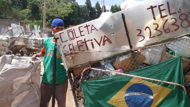 A man standing beside a garbage cart and holding a big sack. The cart has a flag of Brazil and a sign saying "collective collection" on Portuguese.