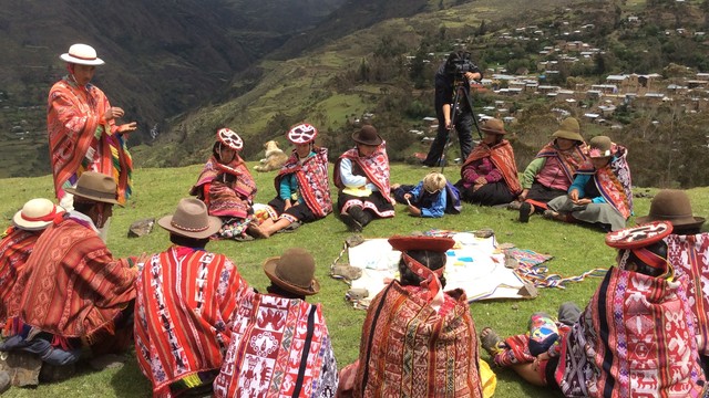 People wearing traditional clothing sit in a circle in a field.