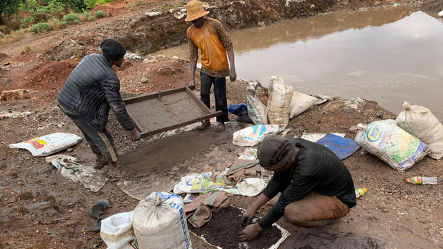 Two men carry a tray on the muddy side of a stretch of water, while another man, bending down, sorts through small stones
