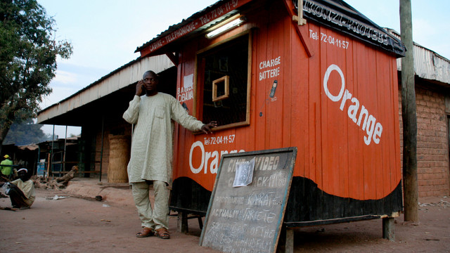 A man on the phone outside a small orange hut. A nearby sign advertises prices for mobile phone air-time.