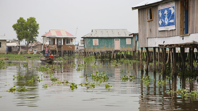 Two people sail a canoe around stilt houses.
